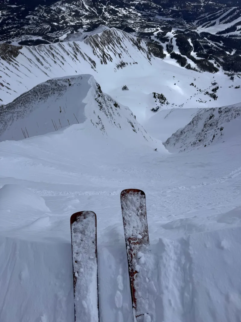 Ski tips at the top of a steep chute, looking down to the lifts below